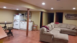 Ein Wohnzimmer mit beiger Couch, einem kleinen Esstisch, einem Kühlschrank und einer Mülltonne.