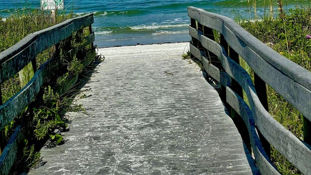 Une promenade en bois mène à une plage où l’eau est d’un bleu cristallin, et on aperçoit un panneau au loin.
