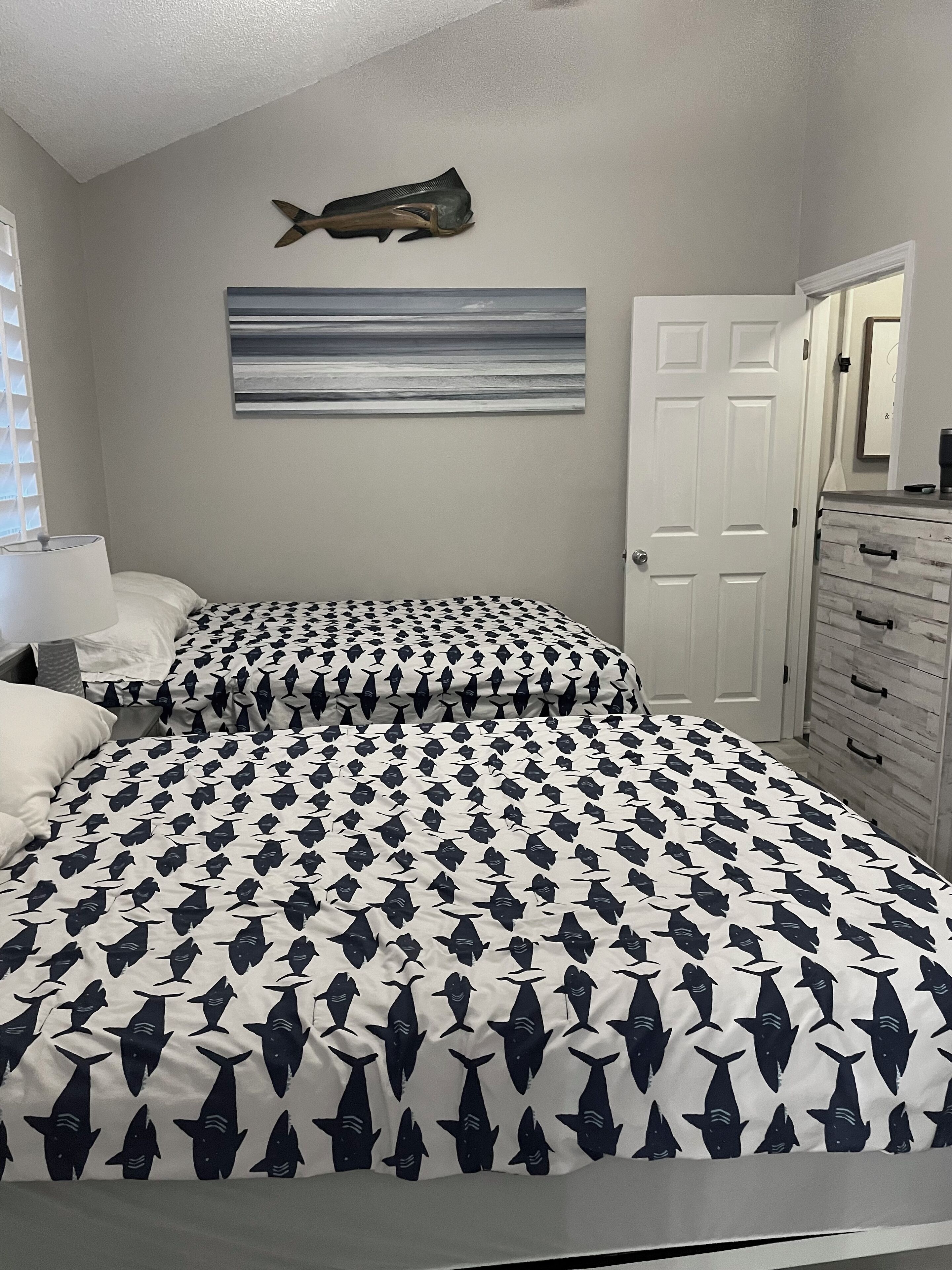 A bedroom with two beds, a fish-patterned bedspread, a wall-mounted fish decoration, a door, and a dresser with drawers.