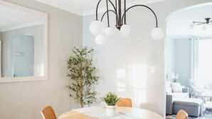 Een eetgedeelte met een ronde tafel, vier stoelen, een kroonluchter, een potplant en een grote spiegel aan de muur.