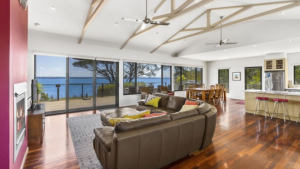 A spacious living area with a large sofa, dining table, and chairs, a kitchen area with bar stools, and large windows offering a view of the sea.