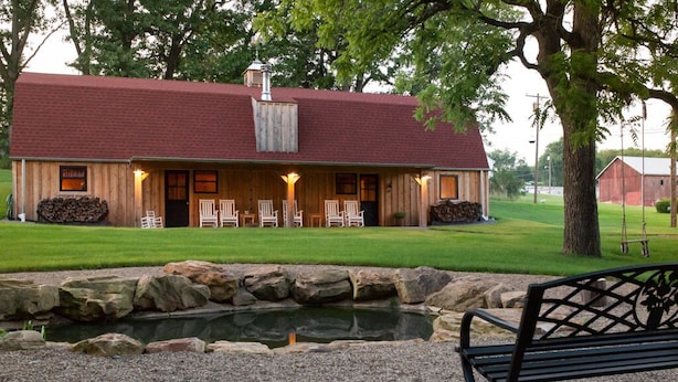 A rustic lodge with a red roof, a covered porch with chairs, a pond, and a bench. - Unique 3 bedroom barn with a fireplace and pond (Sarver)