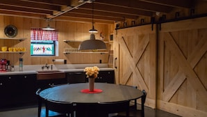 A rustic kitchen with wooden walls, a round table with chairs, a hanging pendant light, and a sliding barn door. - Unique 3 bedroom barn with a fireplace and pond (Sarver)