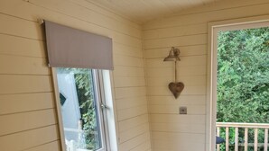 A room with a black leather sofa, a window with a beige blind, a wall-mounted light fixture, and a view of a wooden deck and greenery outside. - Glamping Hut - Sleeps 2, hot tub & parking (Welshpool)