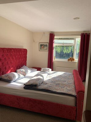 A room with a red upholstered bed, patterned bedding, a window with curtains, and a radiator.