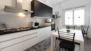Een moderne keuken met witte kasten, een zwart aanrecht en een gasfornuis. Er is een eettafel met stoelen en een raam met witte gordijnen.