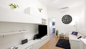 Een moderne woonkamer met een flatscreen televisie, witte meubels, een zwarte klok en een klein plantje op de planken.