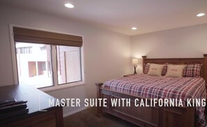 Une suite principale dotée d’un très grand lit (type « California King »), d’une tête de lit en bois, de deux tables de chevet avec lampes et d’une fenêtre avec stores.