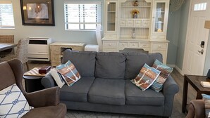 Een woonkamer met een grijze bank, rieten stoelen, een houten salontafel, een witte airconditioner, een witte kast met lades en een lijst met een afbeelding aan de muur.