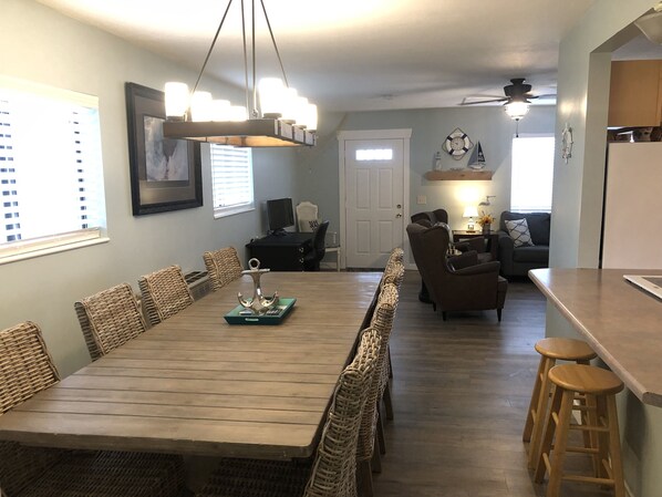 A dining area with a long wooden table surrounded by wicker chairs, a ceiling fan, and a kitchen area with stools and a refrigerator.
