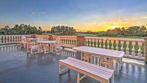An outdoor patio area with wooden tables and benches, a balustrade, and a view of trees and a sunset. - Legends Golf Resort Condo, 4 Beds, 2 Bath Pool access near Coastal Carolina Univ (Myrtle Beach)