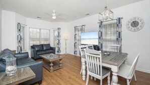 A living room with a sofa, coffee table, and dining table with chairs. There is a TV on the wall and a clock. - Legends Golf Resort Condo, 4 Beds, 2 Bath Pool access near Coastal Carolina Univ (Myrtle Beach)
