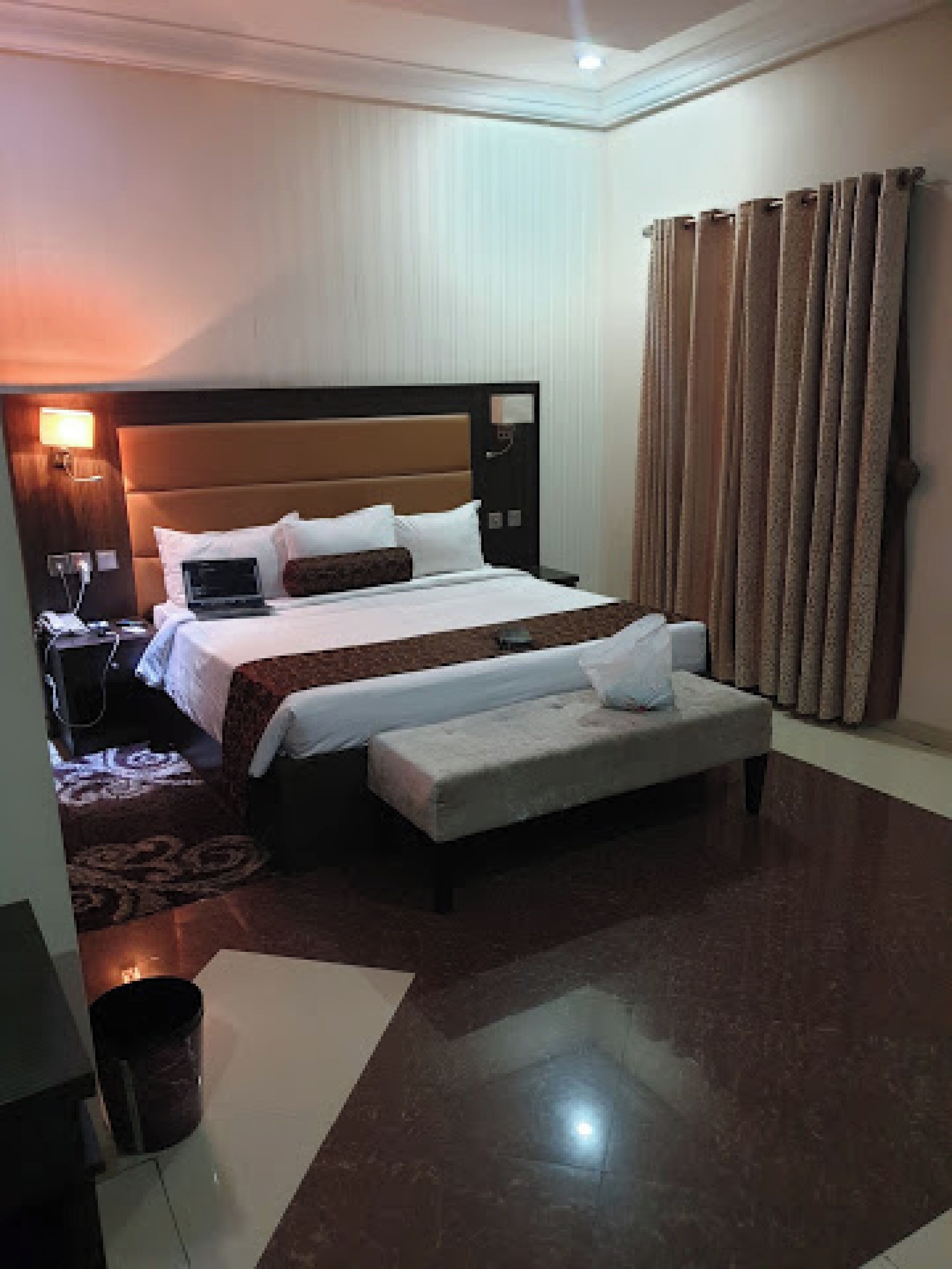 A hotel room with a large bed, a bedside table with a lamp, a laptop, a small table, and a window with curtains.