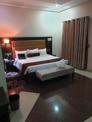Une chambre d’hôtel avec un grand lit, une table de chevet avec une lampe, un ordinateur portable, une petite table et une fenêtre avec des rideaux.