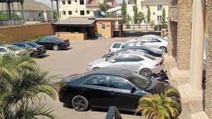 A parking lot with several cars, a building with a red roof, and a stone pillar. - Room-nippon Grand Hotels - Apartment And (Abuja)
