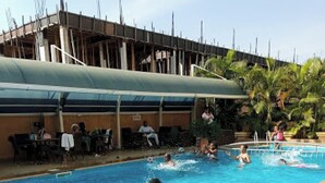 A swimming pool with people in the water, a covered seating area, and a building under construction in the background. - Room-nippon Grand Hotels - Apartment And (Abuja)