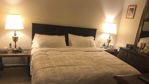 Una cama bien tendida con sábanas blancas, dos lámparas de noche, una mesita de noche con un teléfono, libros y una pequeña mesa con una lámpara.