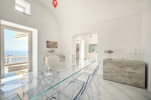 Een moderne, ruime kamer met een glazen eettafel, transparante stoelen, een marmeren kast en uitzicht op zee.