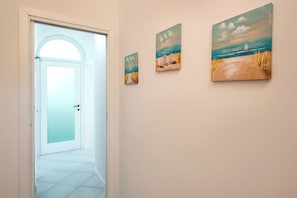 Een hal met een witte deur die naar een kamer leidt, en drie strandafbeeldingen aan de muur.