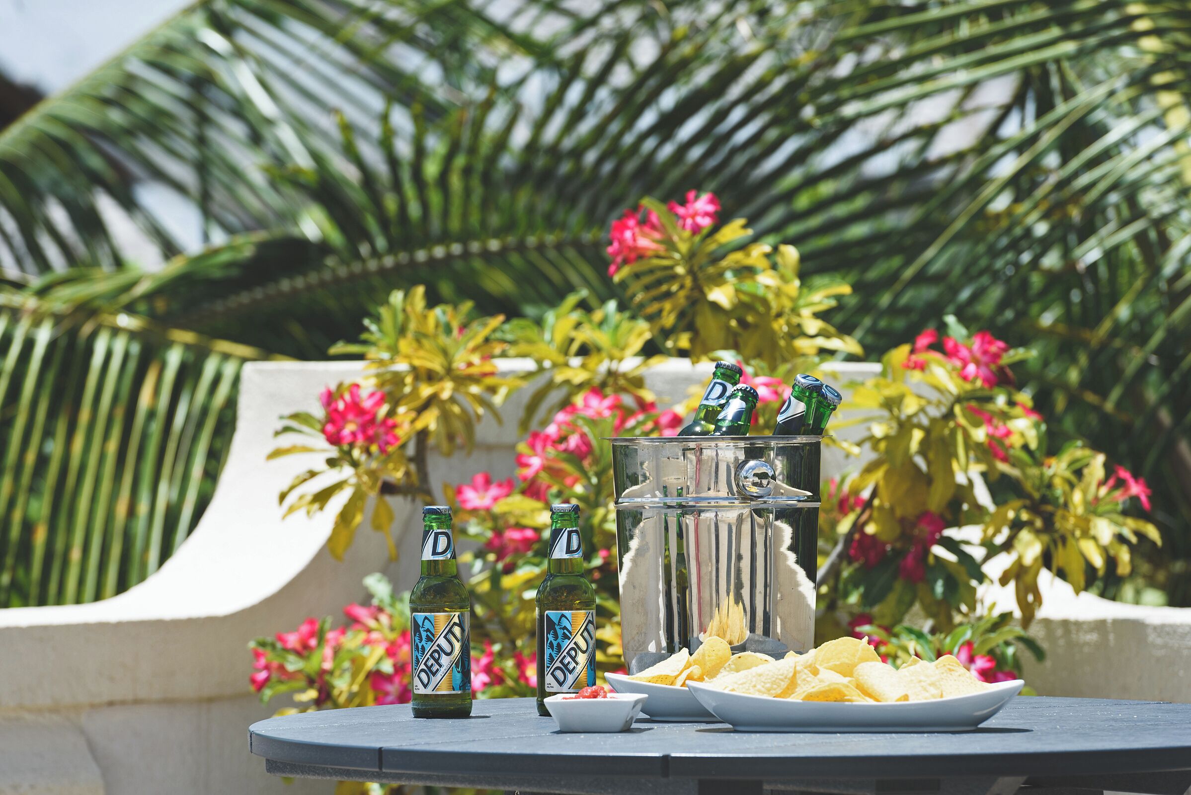 A table with two bottles of beer, a plate of chips, and a large ice bucket with a beverage dispenser, set against a backdrop of lush greenery and pink flowers.
