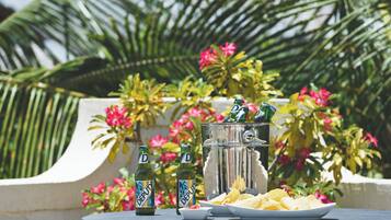 A table with two bottles of beer, a plate of chips, and a large ice bucket with a beverage dispenser, set against a backdrop of lush greenery and pink flowers.