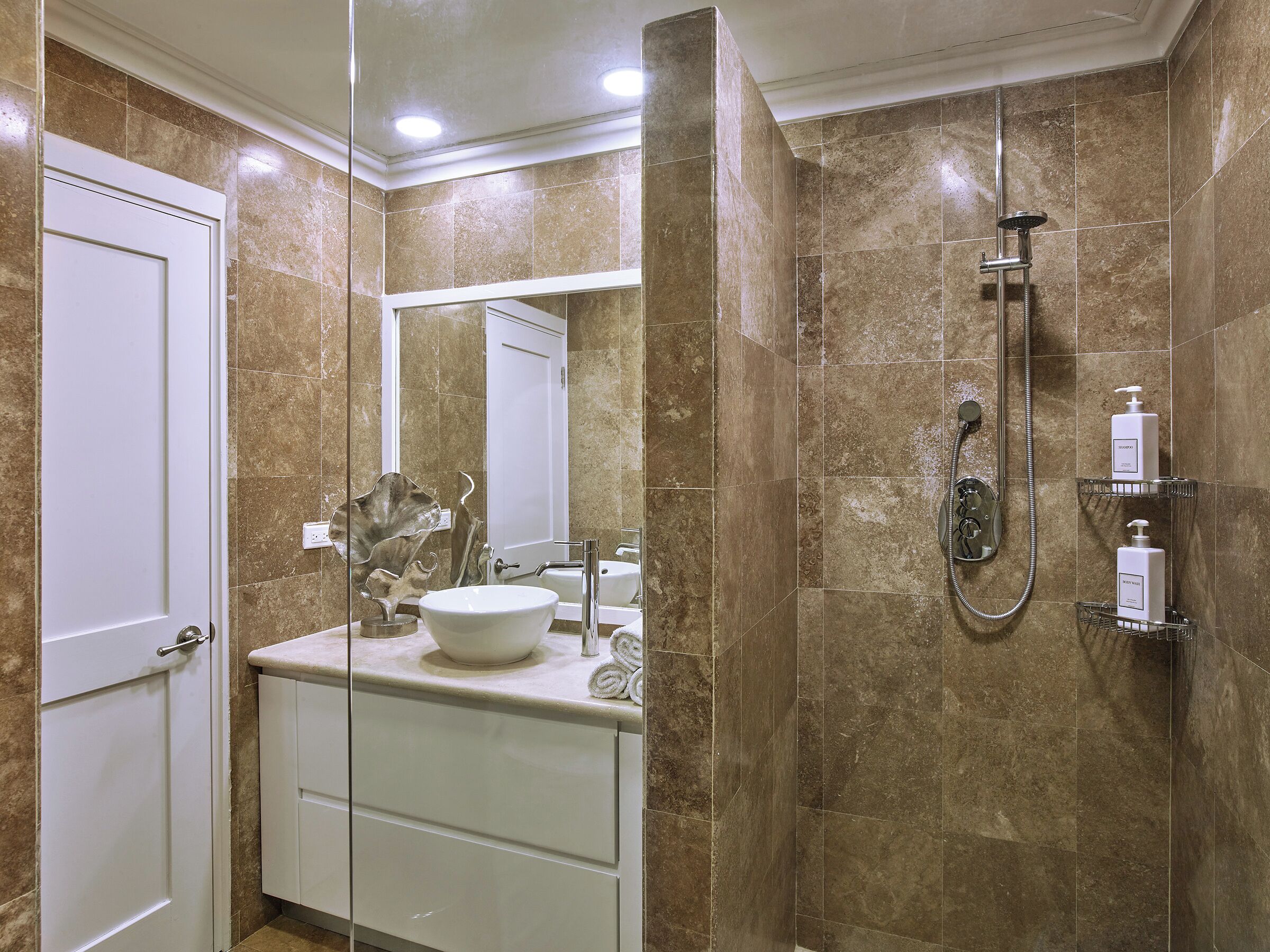 Un baño con lavamanos blanco, un espejo, ducha con cabezal de ducha manual y un perchero para toallas.
