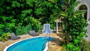 Una piscina con plataforma de baldosas, rodeada de abundante vegetación y un pabellón.