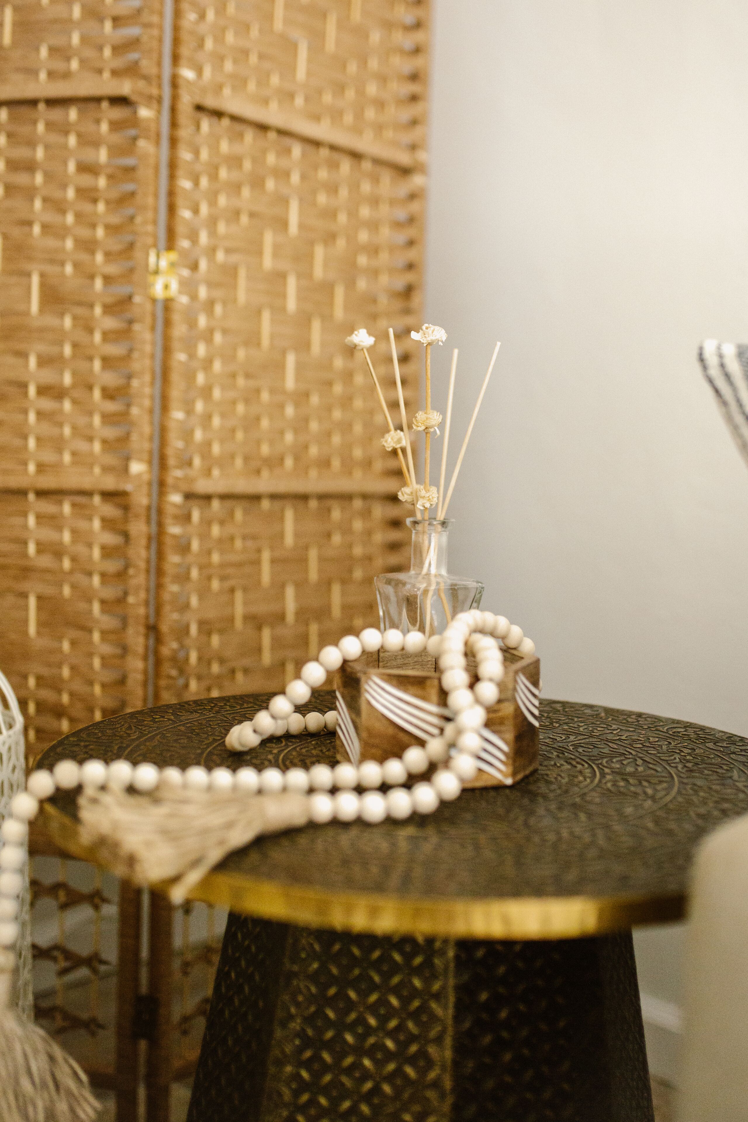 A round table with a decorative glass vase containing a string of white beads and reed sticks.