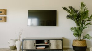 Un soggiorno con una televisione a schermo piatto montata a parete, un mobiletto in legno con ripiani aperti e un'ampia pianta in vaso.