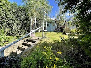 Un jardin avec un escalier en bois menant à une structure blanche, entouré de verdure et sous un ciel dégagé.