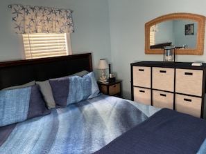 A bedroom with a bed, bedside table, dresser, window with blinds, and a mirror.