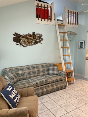 A living room with a plaid sofa, a brown armchair, a ladder, and a wall-mounted shelf with various items.