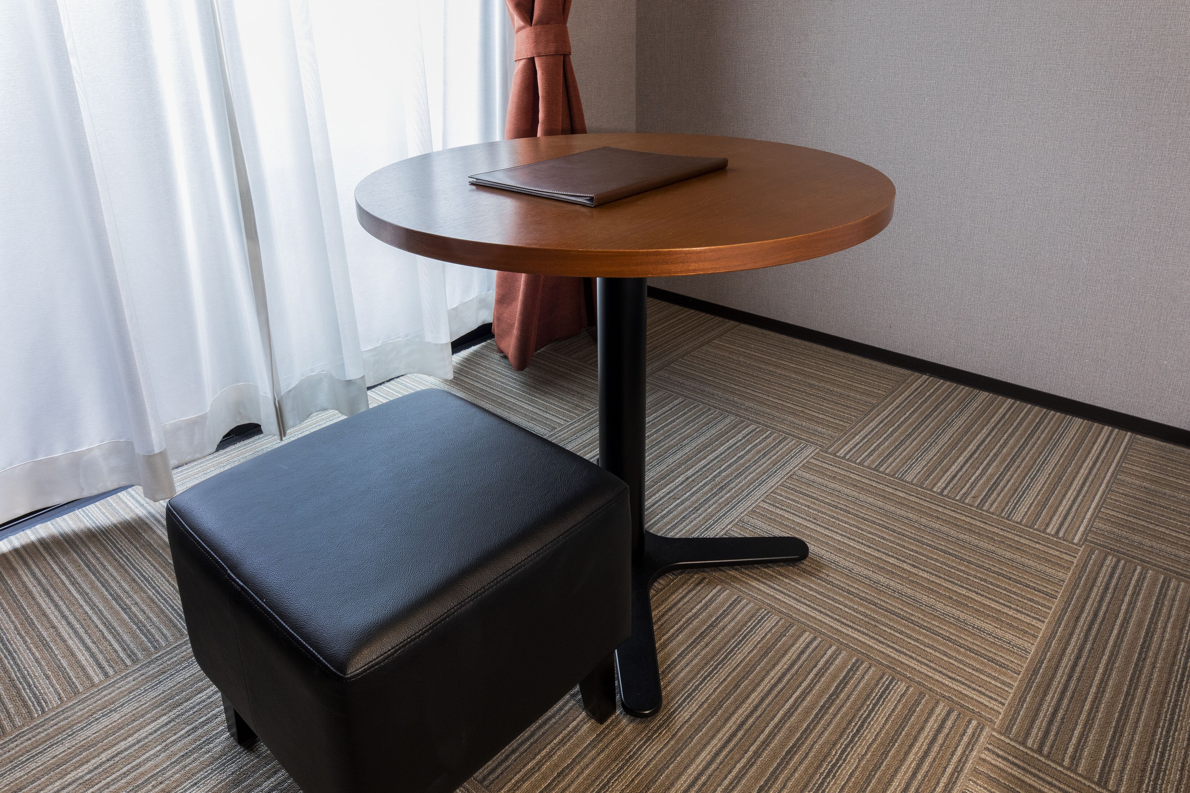 A small, round wooden table with a laptop on top, a black ottoman, and a carpeted floor.