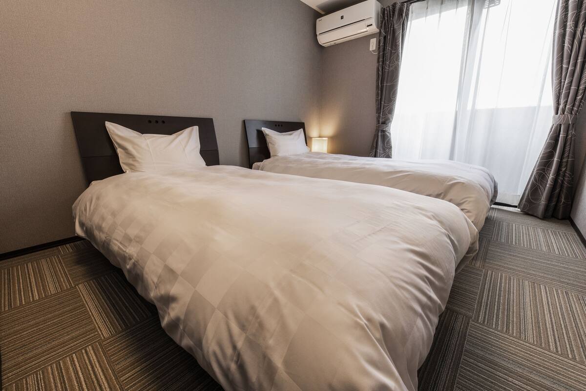 A hotel room with two beds, a headboard, a wall-mounted air conditioner, and a window with curtains.
