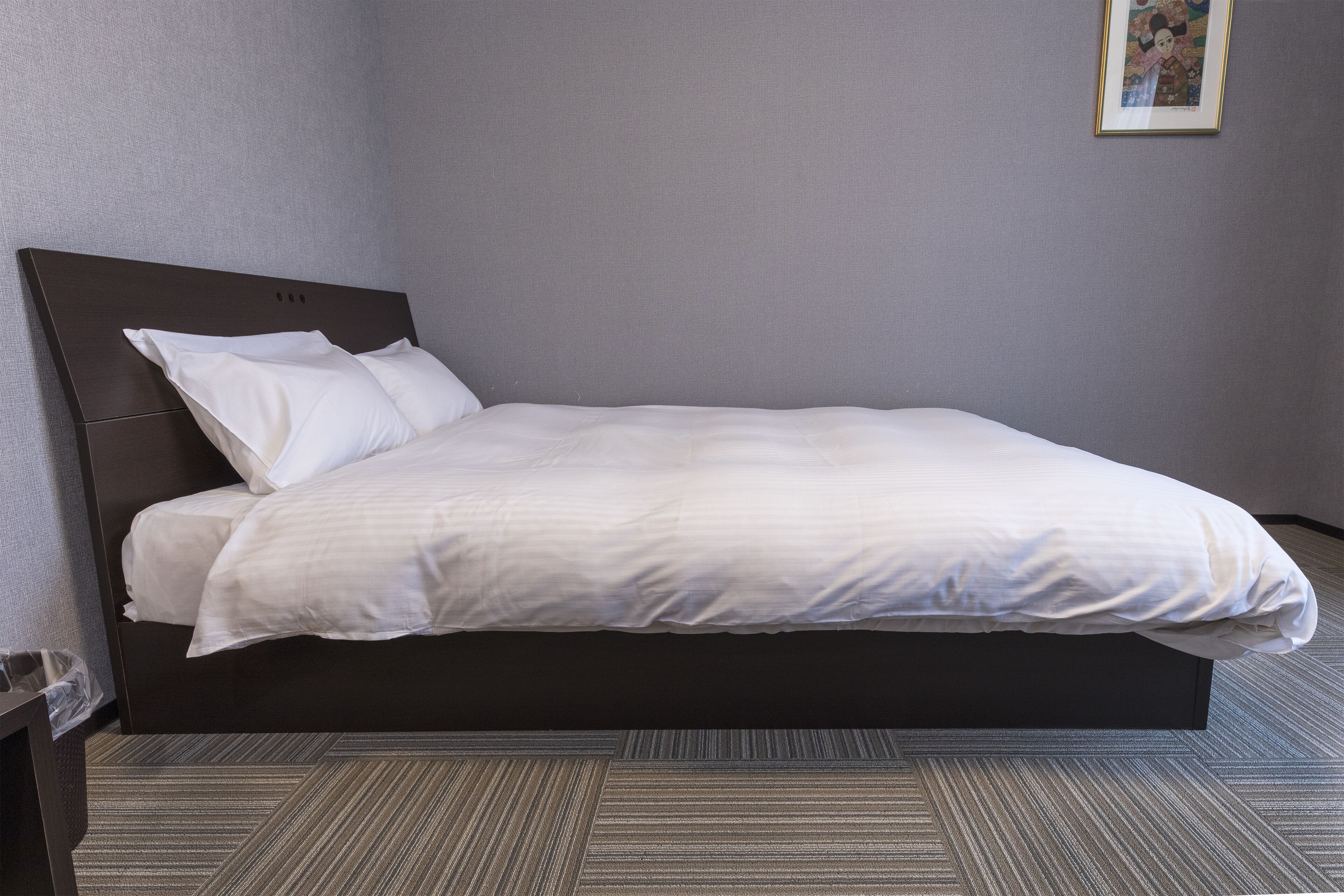 Un lit bien fait dans une chambre d’hôtel, avec une tête de lit sombre, un mur gris et un tableau encadré au-dessus du lit.