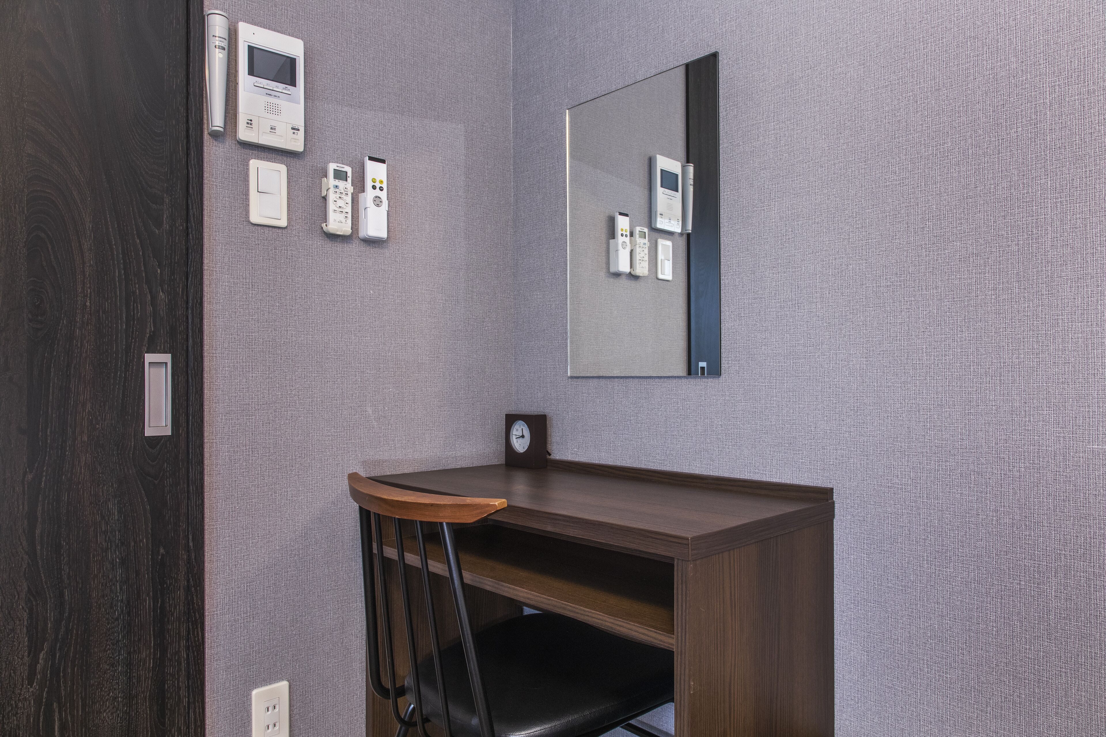 A small, private office with a desk, chair, and a large mirror. There are several electronic devices mounted on the wall, including a thermostat, a phone, and a clock.