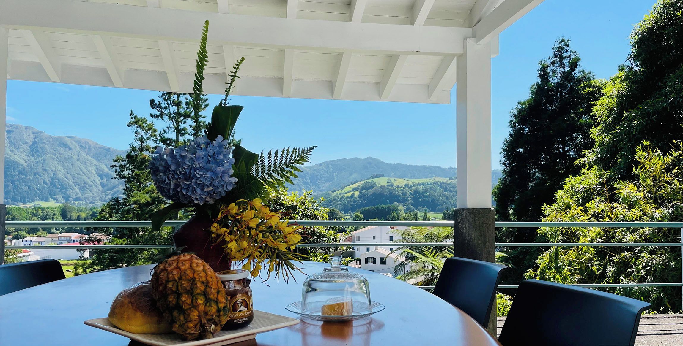Un tavolo con un'ananas, una bottiglia di salsa e una cupola di vetro su un balcone affacciato su una montagna.