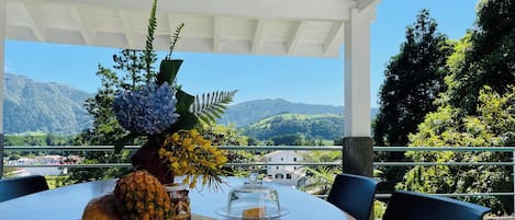 Een tafel met een ananas, een fles saus en een glazen koepel op een balkon met uitzicht op een berg.