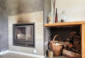 A room with a built-in fireplace, a concrete wall, a wooden shelf with decorative items, and a wicker basket. - Casa de Férias Familiar (Furnas)