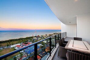 A balcony with wicker furniture overlooking a coastal cityscape with buildings, a beach, and a clear sky.