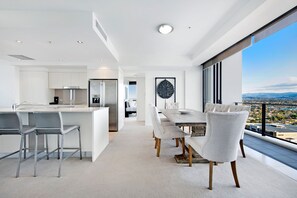 Una cocina moderna con muebles blancos y electrodomésticos de acero inoxidable, un comedor con una mesa grande y sillas, y un balcón con vista a la ciudad.