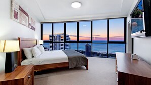 Quarto com uma grande janela que oferece vista para a cidade, uma cama, uma mesinha de cabeceira e uma escrivaninha.