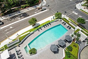 An aerial view of a swimming pool with lounge chairs and umbrellas, surrounded by a landscaped area with trees and a road.