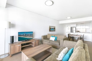 A modern living room with a flat-screen TV, a sofa, a coffee table, and a dining area with a kitchen in the background.