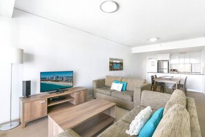 A modern living room with a flat-screen TV, a sofa, a coffee table, and a dining area with a table and chairs. - Hinterland Views 2BR apartment - Sierra Grand (Broadbeach)