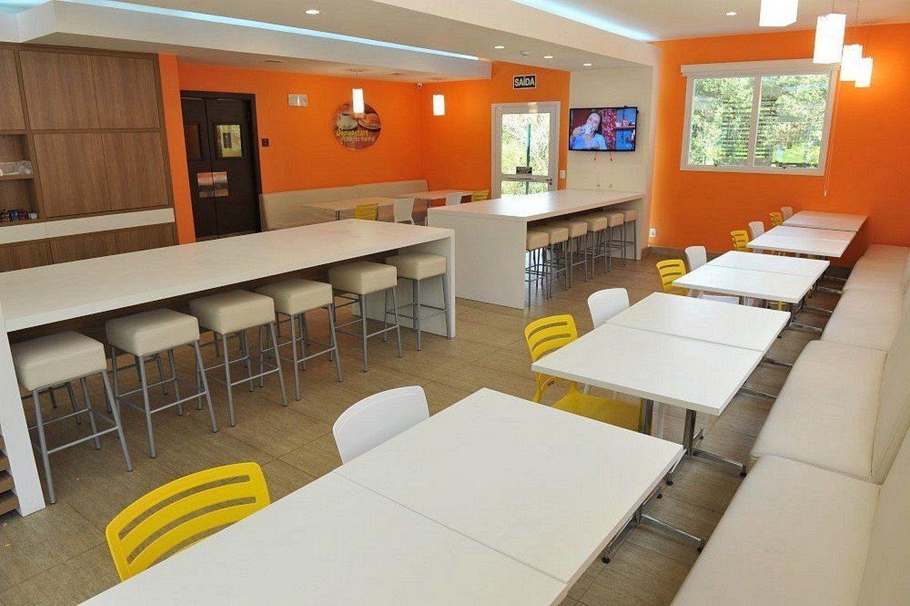 Uma área de jantar moderna com mesas brancas e cadeiras amarelas, paredes laranja e uma TV na parede.