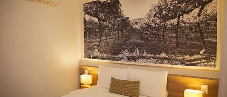 A hotel room with a large bed, two bedside lamps, a wall-mounted air conditioner, and a large black-and-white landscape painting above the bed.