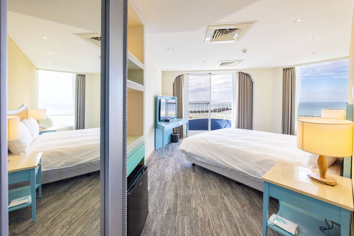 Hotellihuone, jossa on kaksi sänkyä, työpöytä, tuoli, televisio ja näkymä merelle.