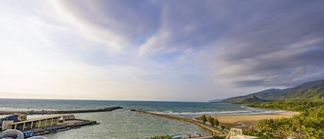 Vista costeira com um cais que se estende para o mar, uma praia e montanhas ao fundo.