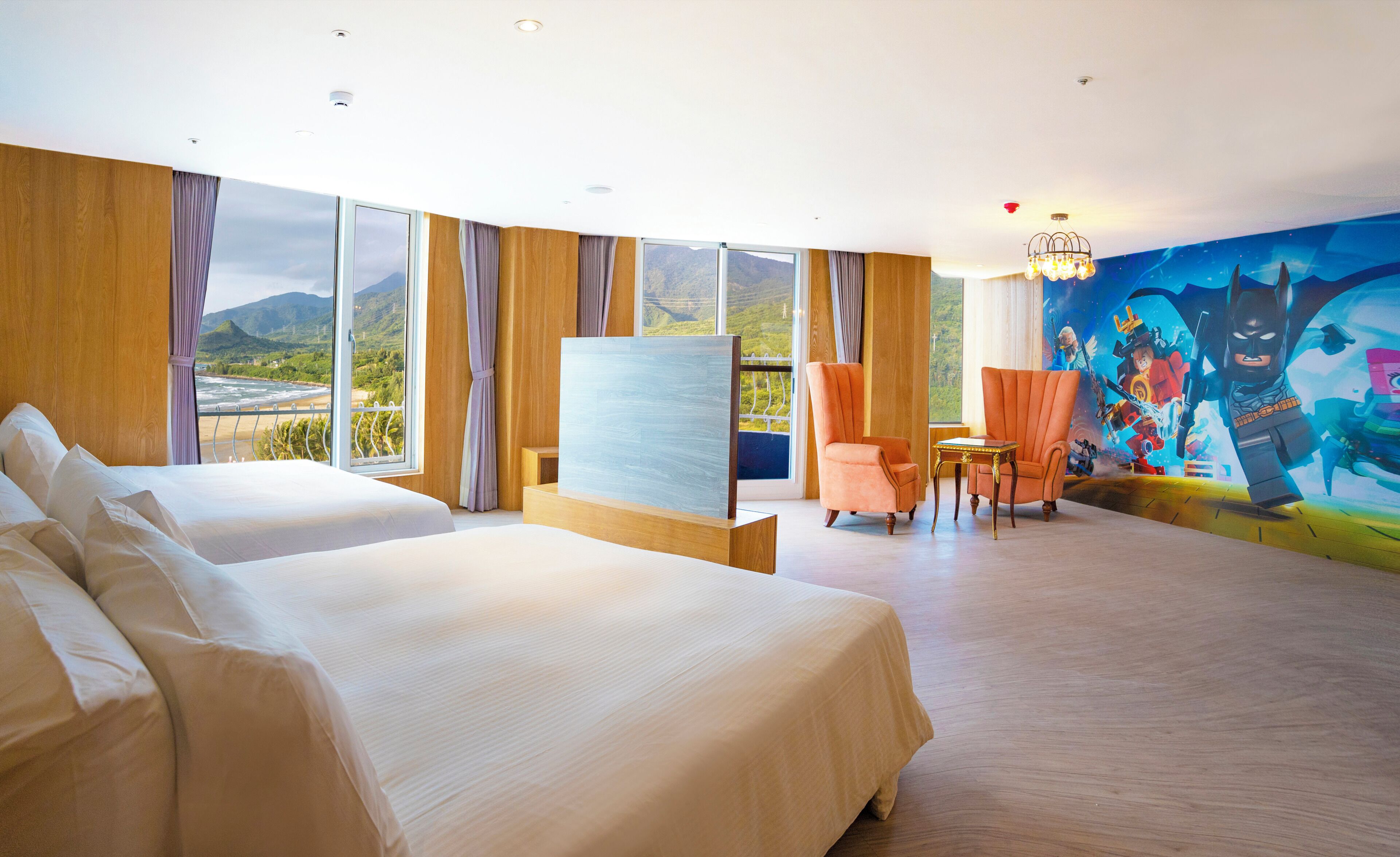A hotel room with two beds, a large window with a view of a mountainous landscape, a painting on the wall, and a small table with a chair.